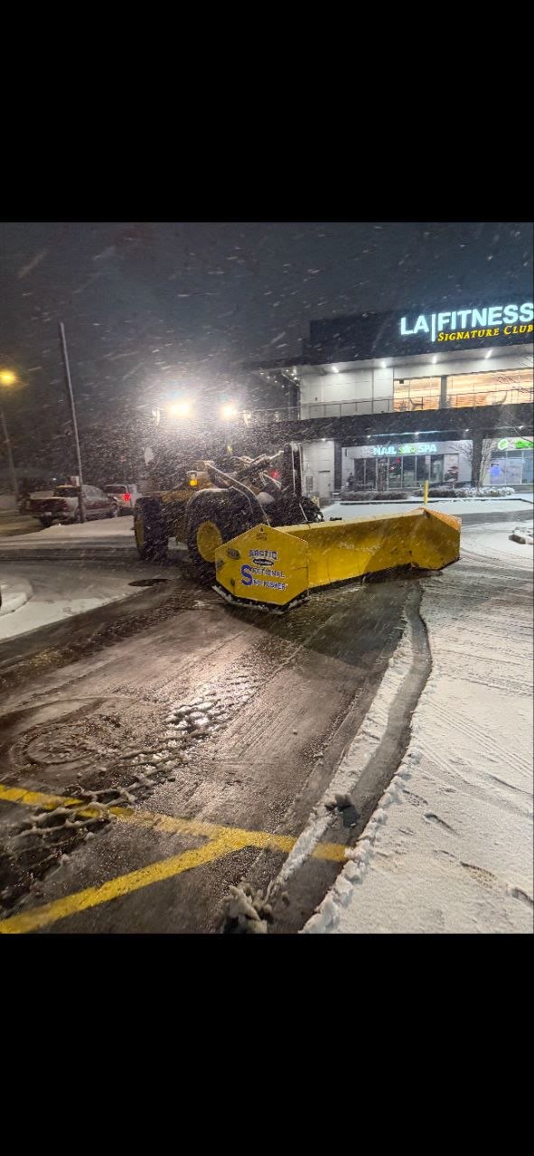 Arctic Sectional Sno-Pusher on a wheel loader clearing a LA Fitness-style retail parking lot at night during a storm